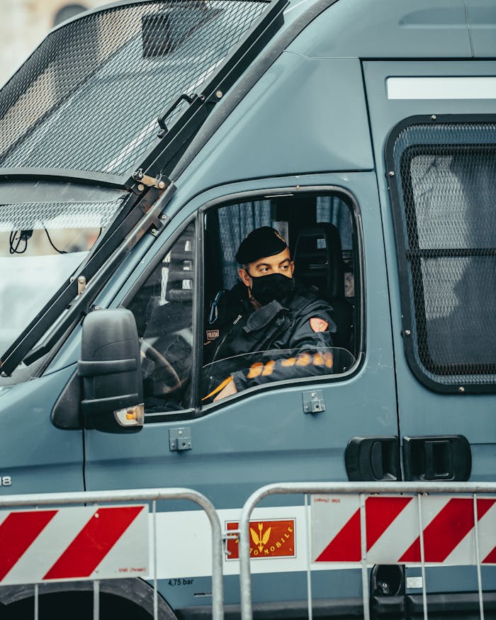 A police officer wearing a mask is seated inside a secure van behind a barricade.